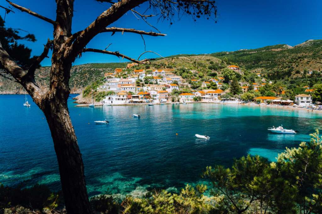 Assos Village, Kefalonia Greece. View On Turquoise Transparent Bay Lagoon Of Mediterranean Sea. Surrounded By Green Pine Trees. Blue Deep Pattern On Bottom